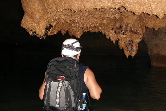 A caver in a white helmet and headlamp, seen from behind, facing stalactites in a dark cave