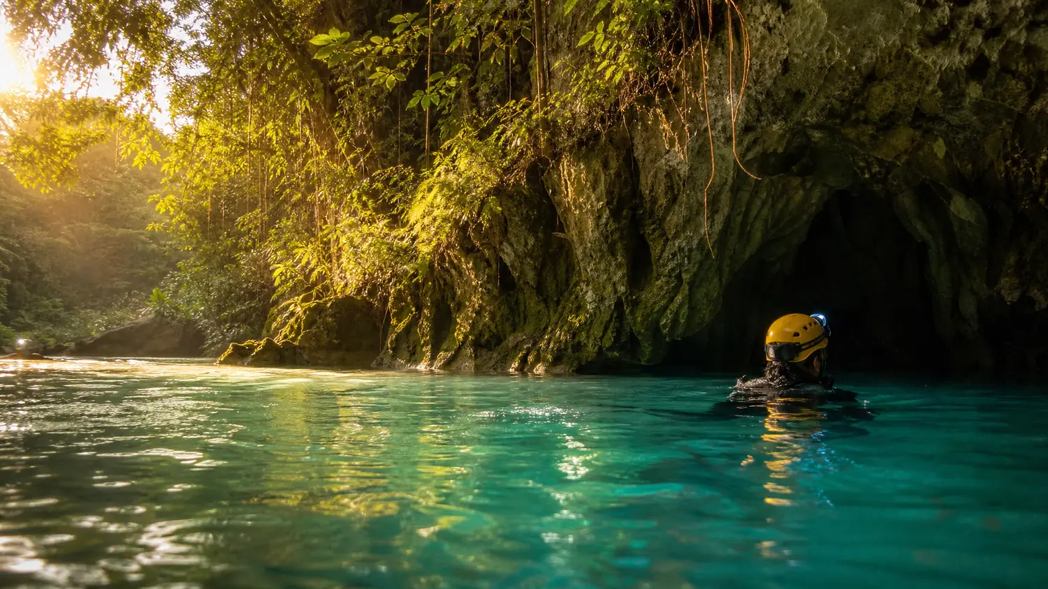 A swimmer in a yellow helmet heading into a cave mouth across green water at golden hour
