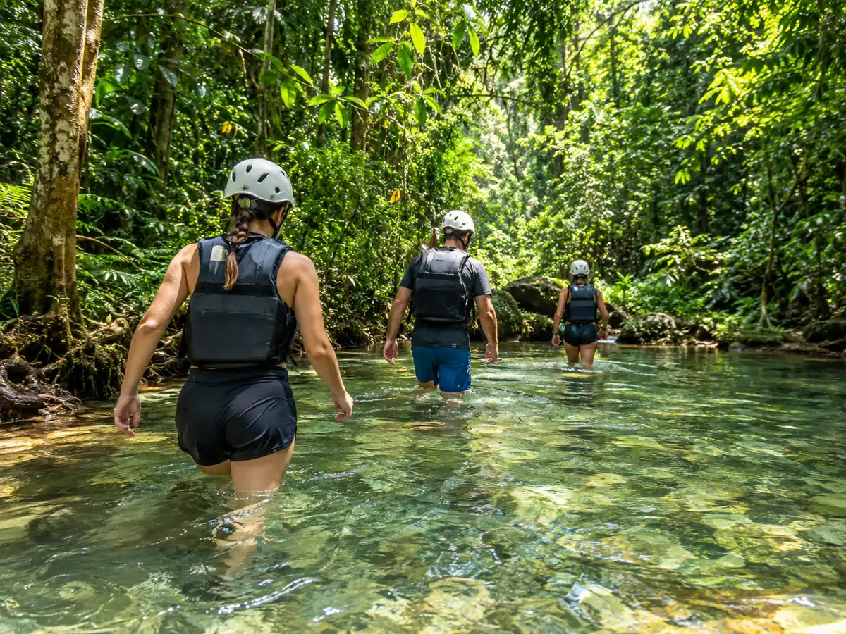 Hikers in helmets and life vests crossing a clear jungle stream